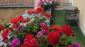 A courtyard with a small table and chairs, surrounded by vibrant geraniums. A large flower pot with pink and red flowers is visible. The background features a building with a tiled roof and a tower with pointed spires.