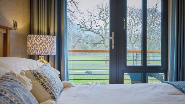 A bedroom with a large bed, a patterned lamp, and a view of a park with a fence and trees. - Riverside Park 3 (Keswick)