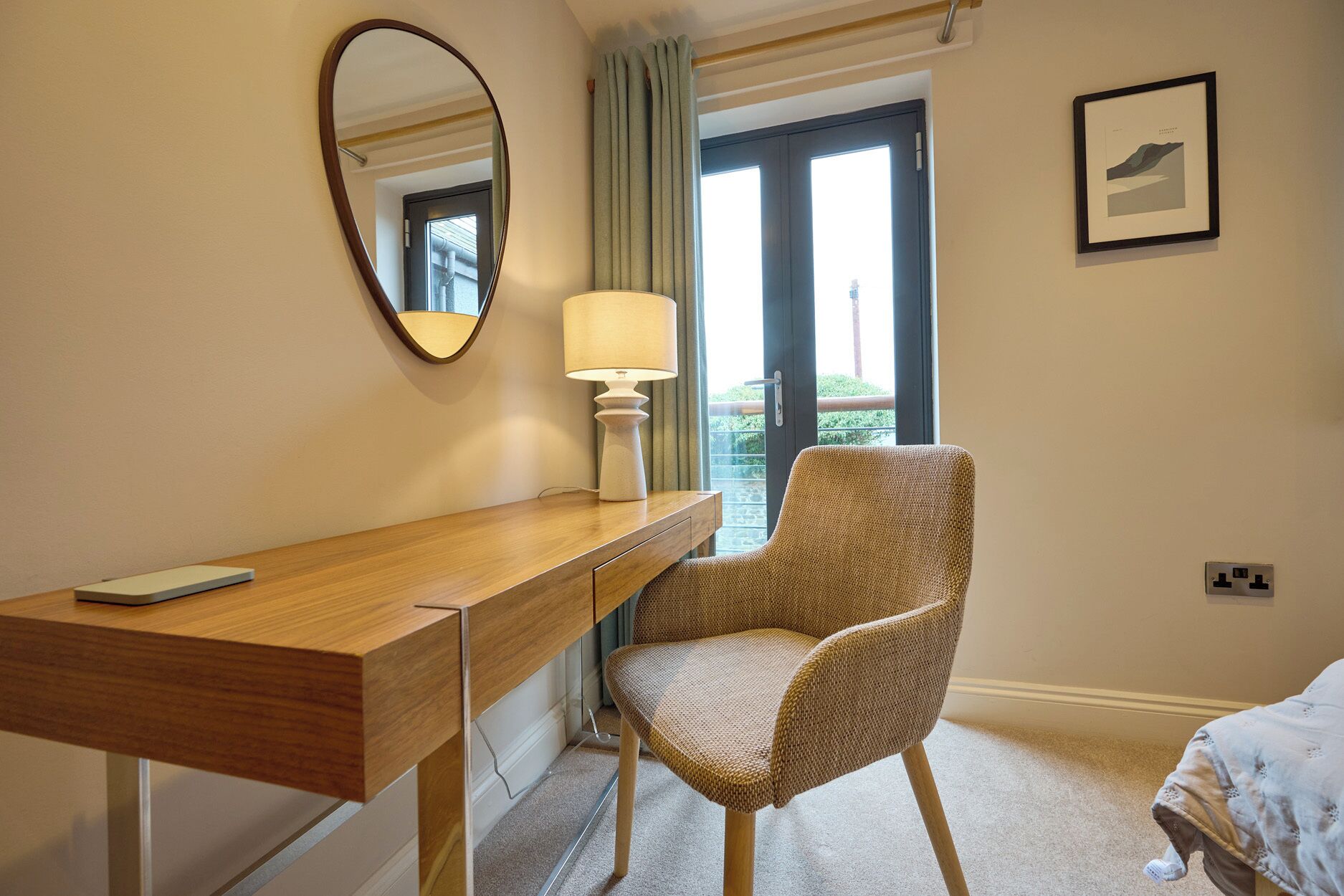 A hotel room with a wooden desk, a wicker chair, a bedside table, a lamp, a bed, a window with curtains, and a framed picture on the wall.