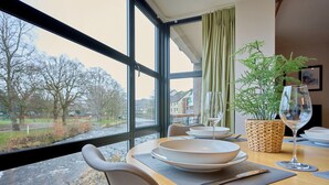 A dining table with plates, cutlery, and a glass set, positioned near a large window with a view of a river and buildings. - Riverside Park 3 (Keswick)
