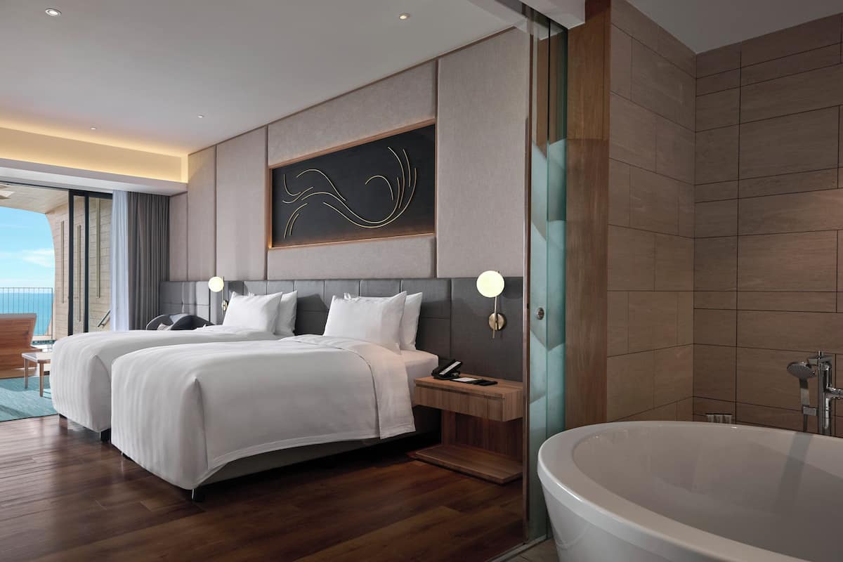 Une chambre d’hôtel moderne dotée d’un grand lit, d’un coin salon séparé, d’une baignoire et offrant une vue sur la mer.
