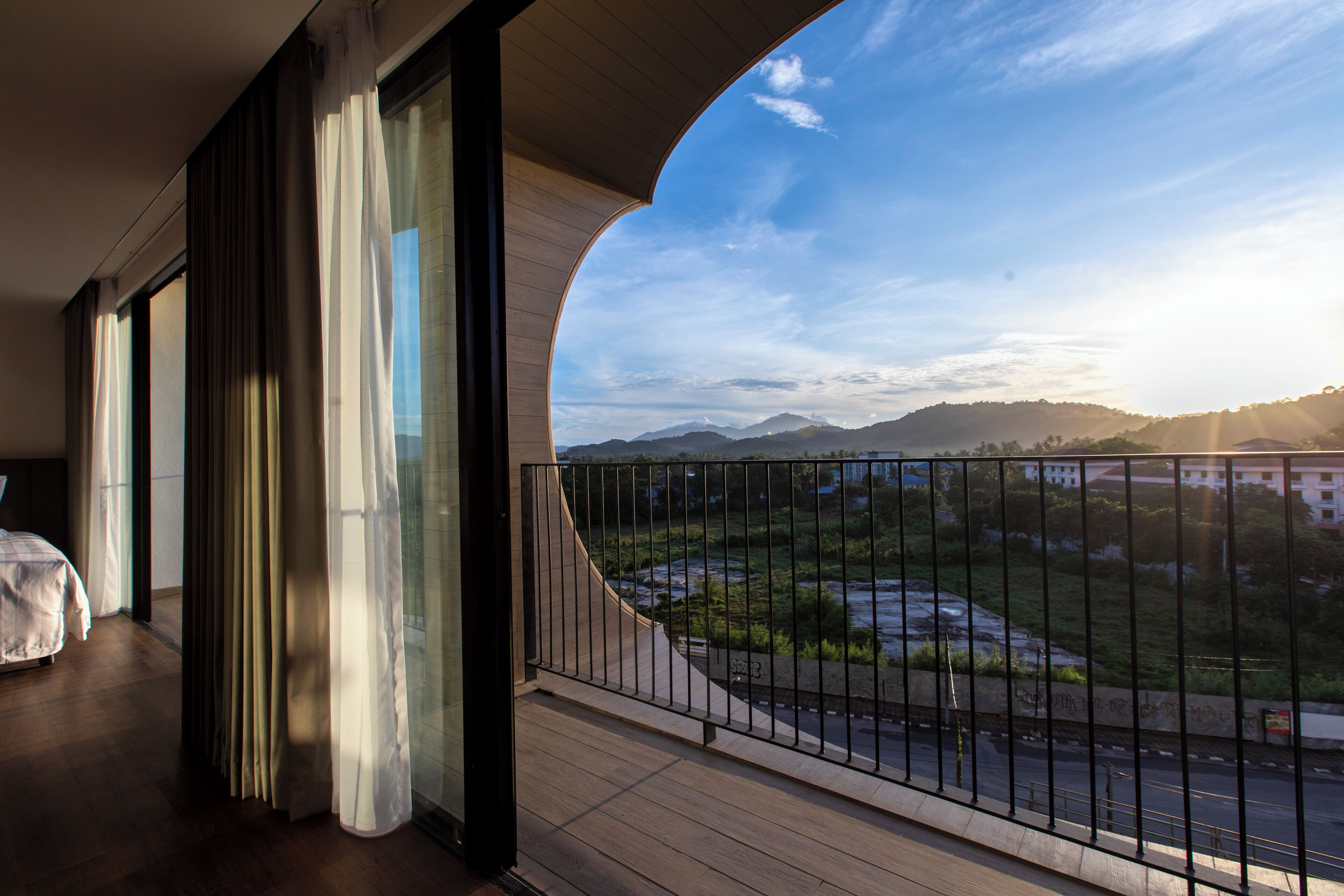 A balcony with a view of a river and mountains, a bed, and a window with curtains.