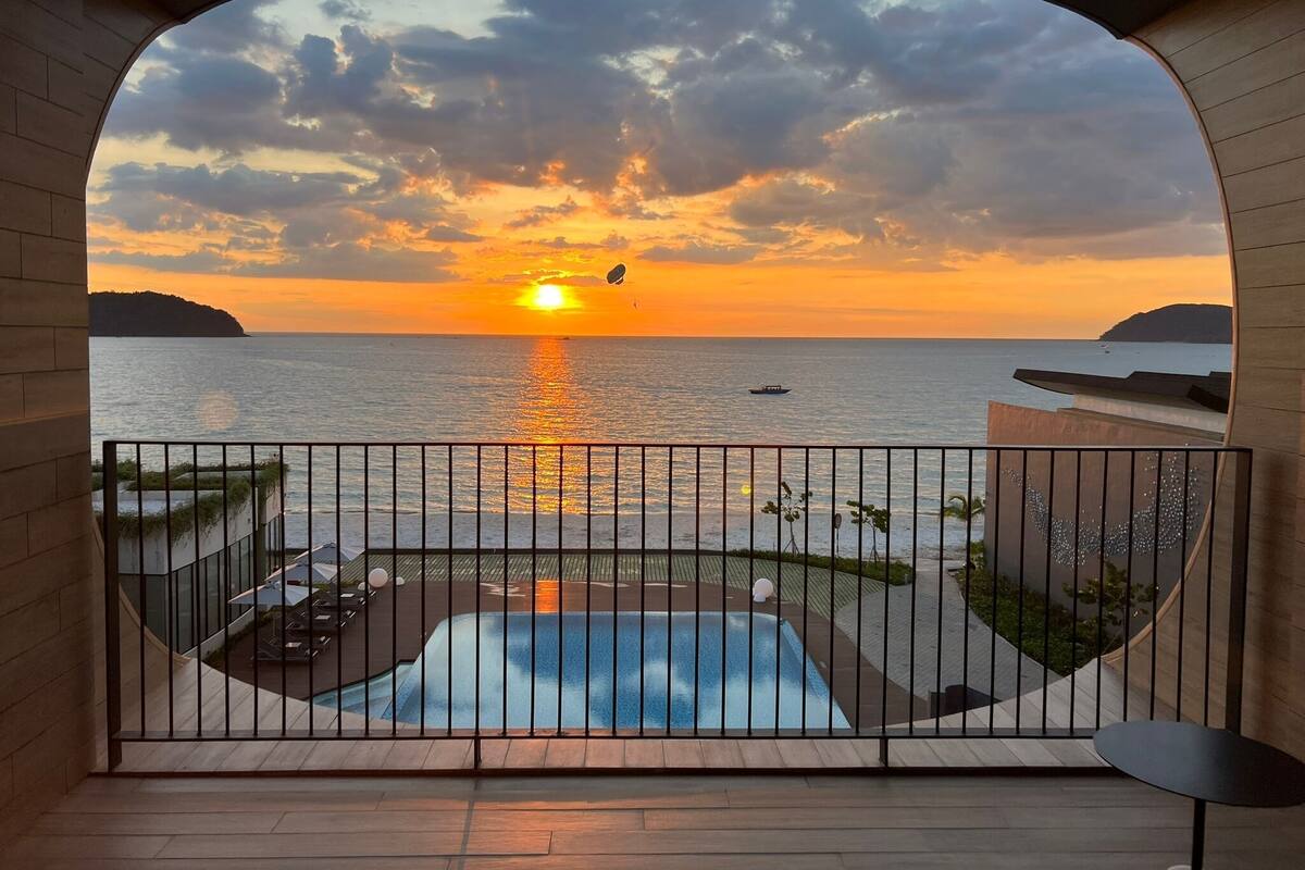 Un balcon avec vue sur le coucher de soleil sur la mer, une piscine et une petite table.