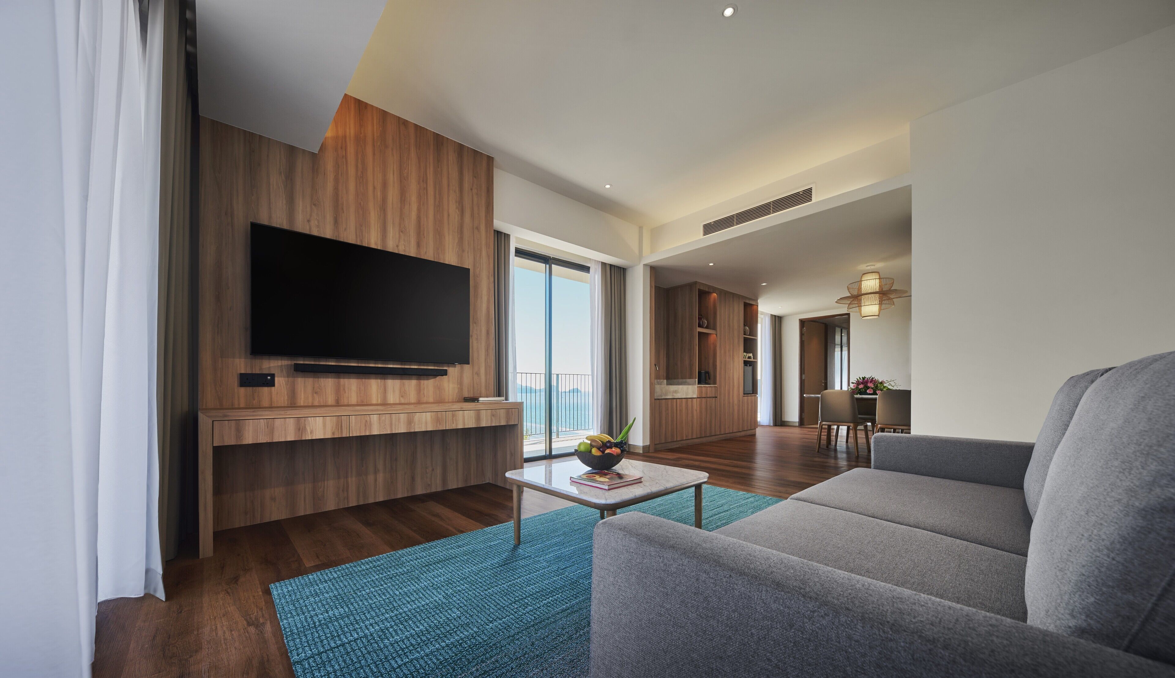 A modern living room with a flat-screen TV, a sofa, a coffee table with a fruit bowl, and a view of the outdoors.