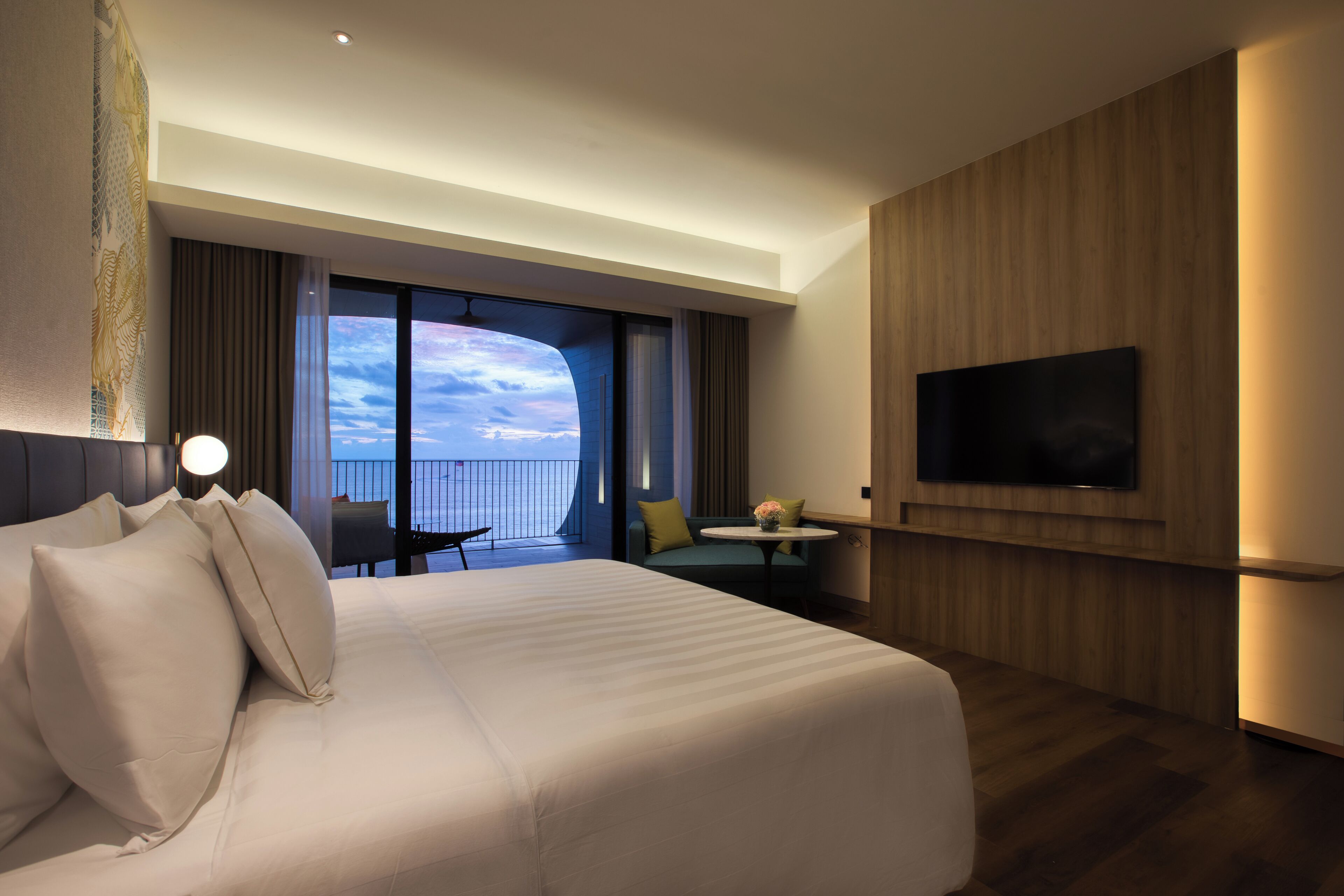 A hotel room with a large bed, a TV mounted on the wall, a balcony with a view, and a seating area with a table.