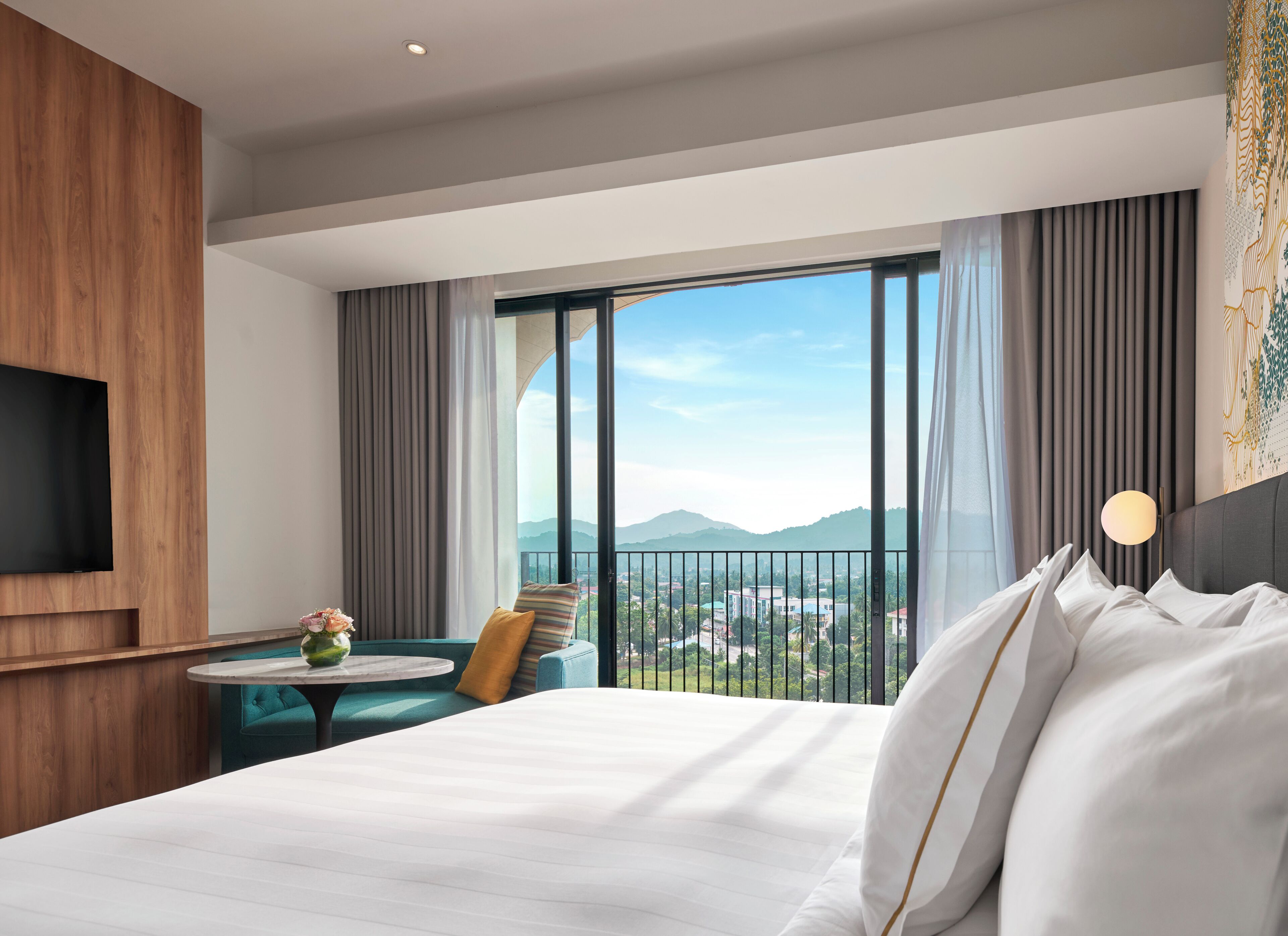 A hotel room with a large bed, a small round table with a vase of flowers, a teal armchair, and a view of the city and mountains through large windows.