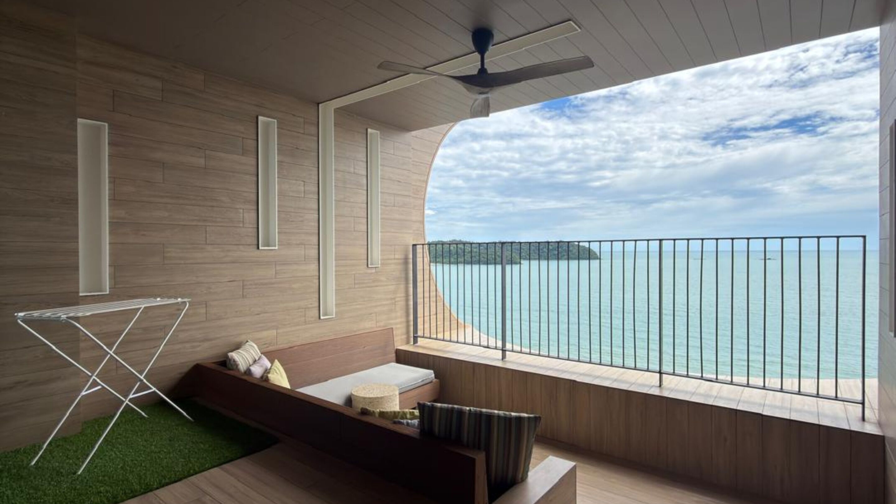 Quarto moderno com piso de madeira, ventilador de teto, janela com vista para o mar e um pequeno espaço de estar com almofadas.