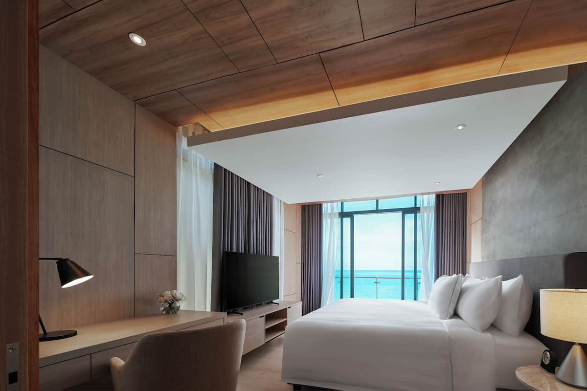 Une chambre d’hôtel moderne avec un grand lit, un bureau équipé d’une télévision et d’une lampe, et une vue sur la mer.