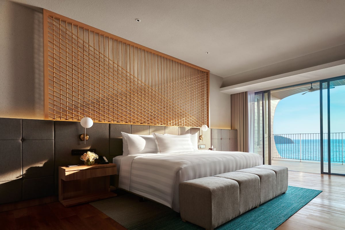 Une chambre d’hôtel moderne dotée d’un grand lit, d’une tête de lit en bois, d’un bureau et offrant une vue sur l’océan.