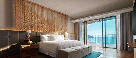 A modern hotel room with a large bed, a wooden headboard, a desk, and a view of the ocean.