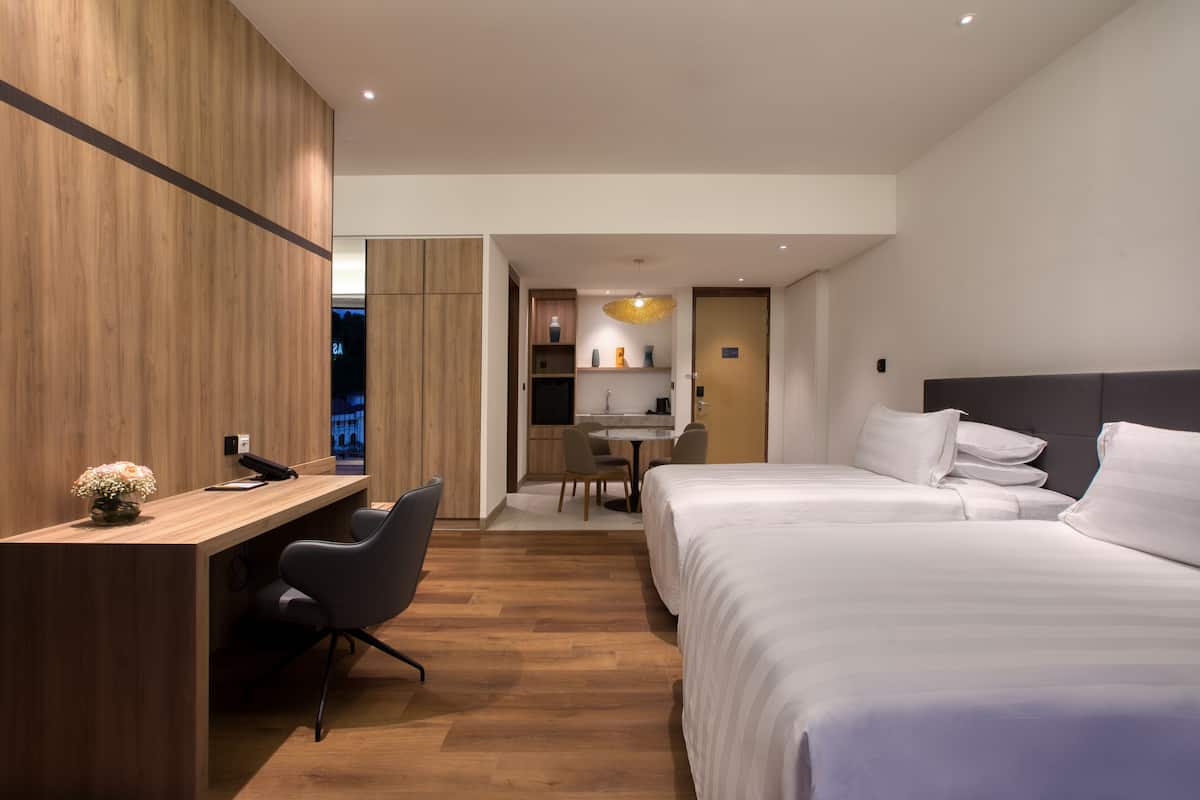 Une chambre d’hôtel moderne avec deux lits, un bureau et une chaise. On aperçoit un coin cuisine au fond de la pièce.