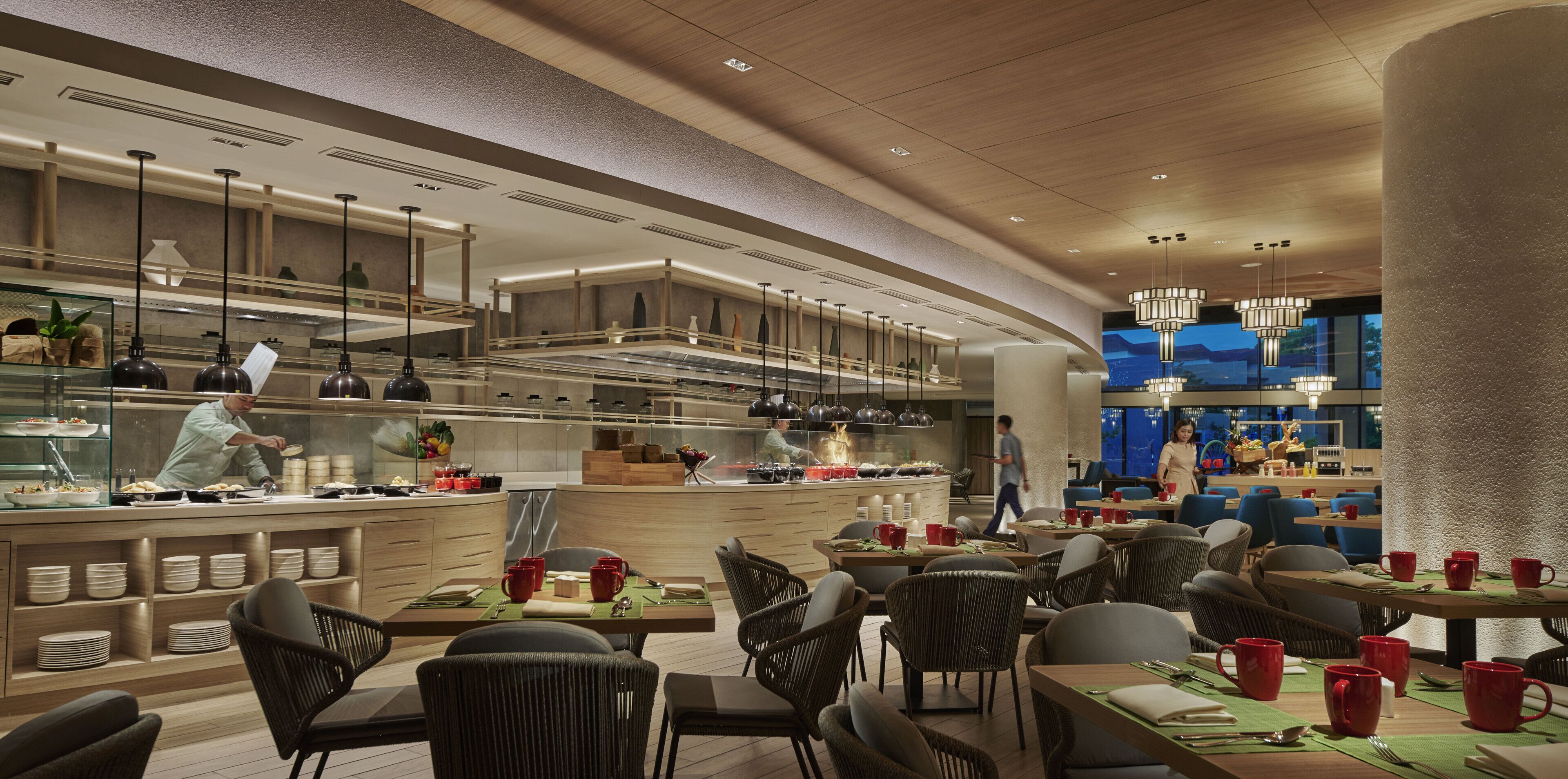 A modern restaurant with a central counter, dining area with wicker chairs, and a bar area with a view of the cityscape.