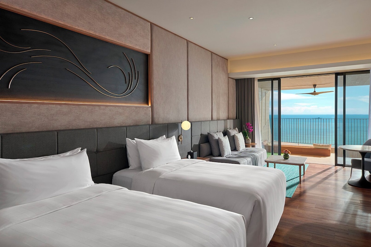 A hotel room with two beds, a sofa, a balcony with a view of the sea, and a modern decor style.