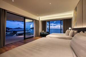 A modern hotel room with two beds, a balcony with a view of the sea, and a dining area with a table and chairs.