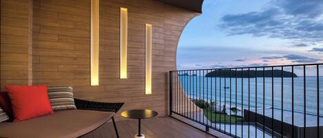 A balcony with a wooden floor, a cushioned seating area, and a view of the sea.