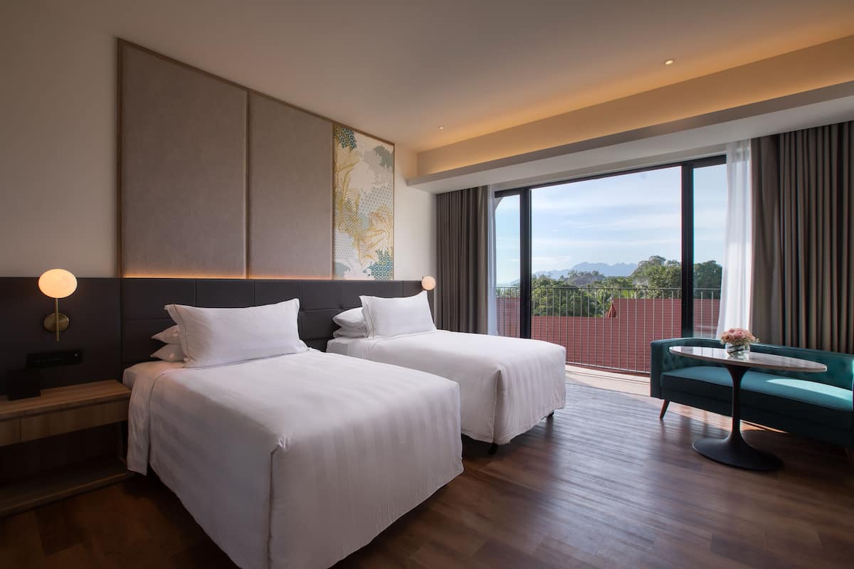 Une chambre d’hôtel avec deux lits, un bureau, une chaise et une grande fenêtre donnant sur l’extérieur.