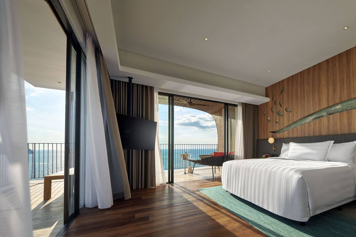 Une chambre d’hôtel moderne dotée d’un grand lit, d’une table de chevet, d’une télévision et d’un balcon offrant une vue sur la mer.