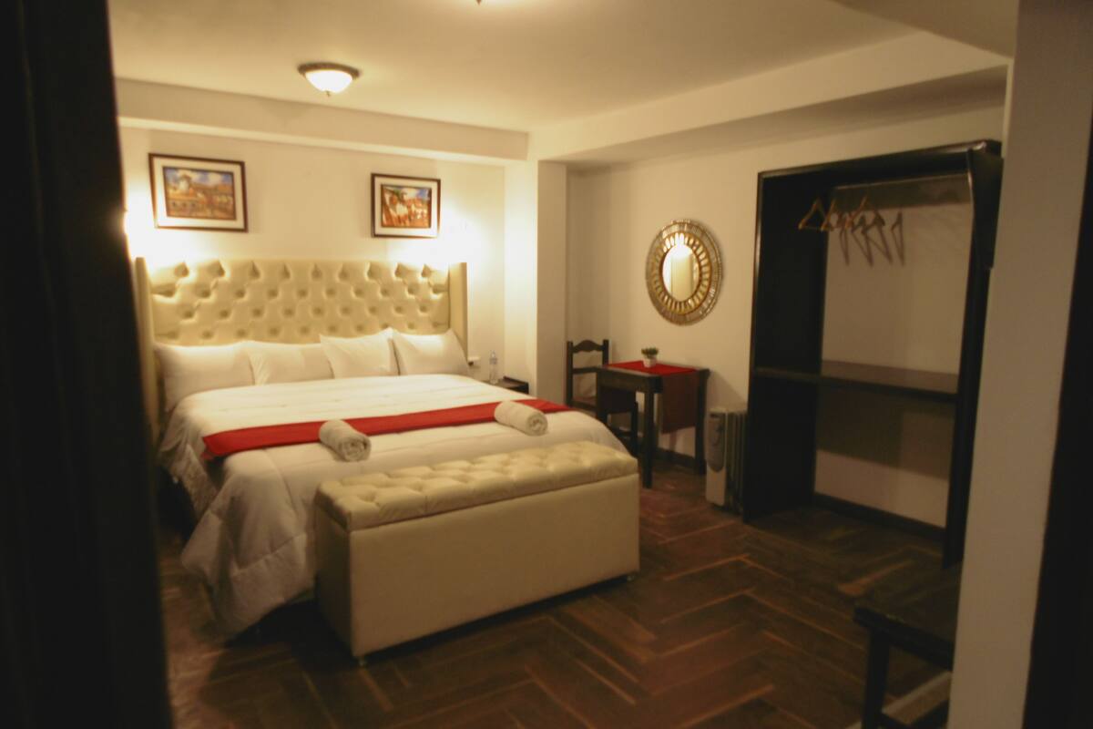 A hotel room with a large bed, a small table, a chair, and a mirror on the wall.