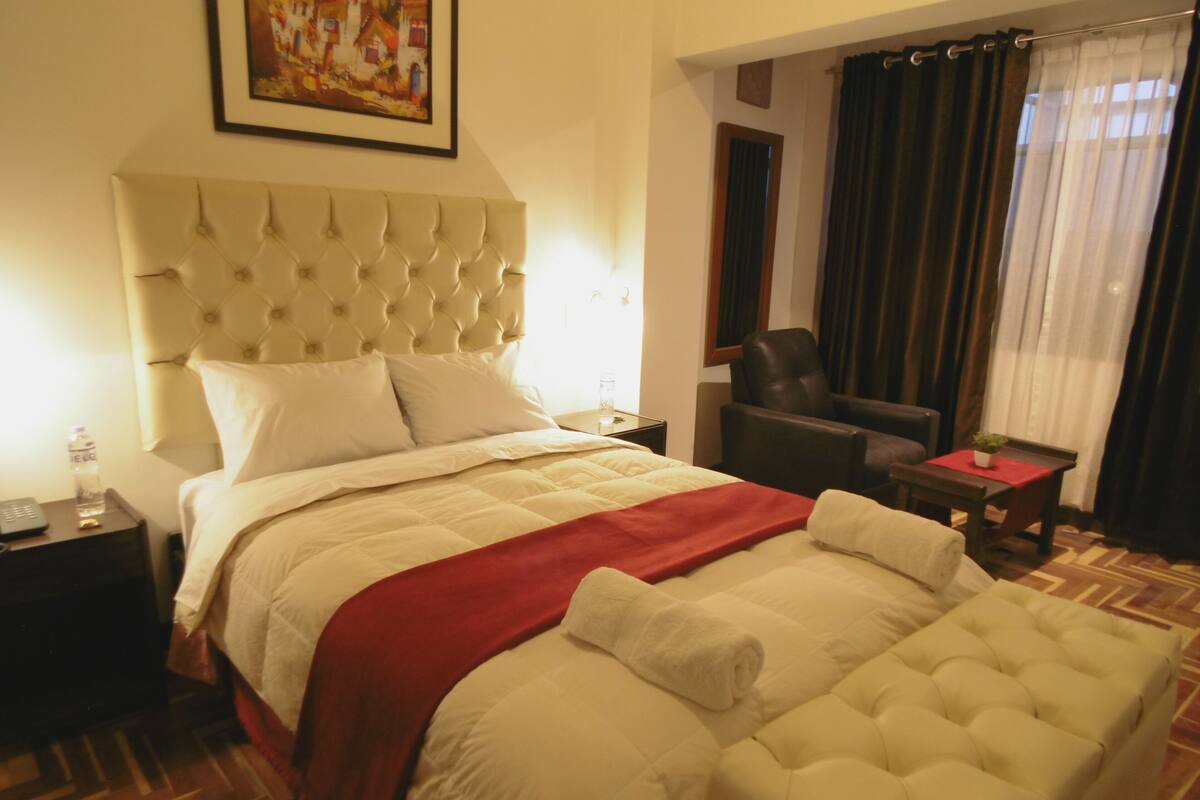 A hotel room with a large bed, a chair, a small table, and a painting on the wall.