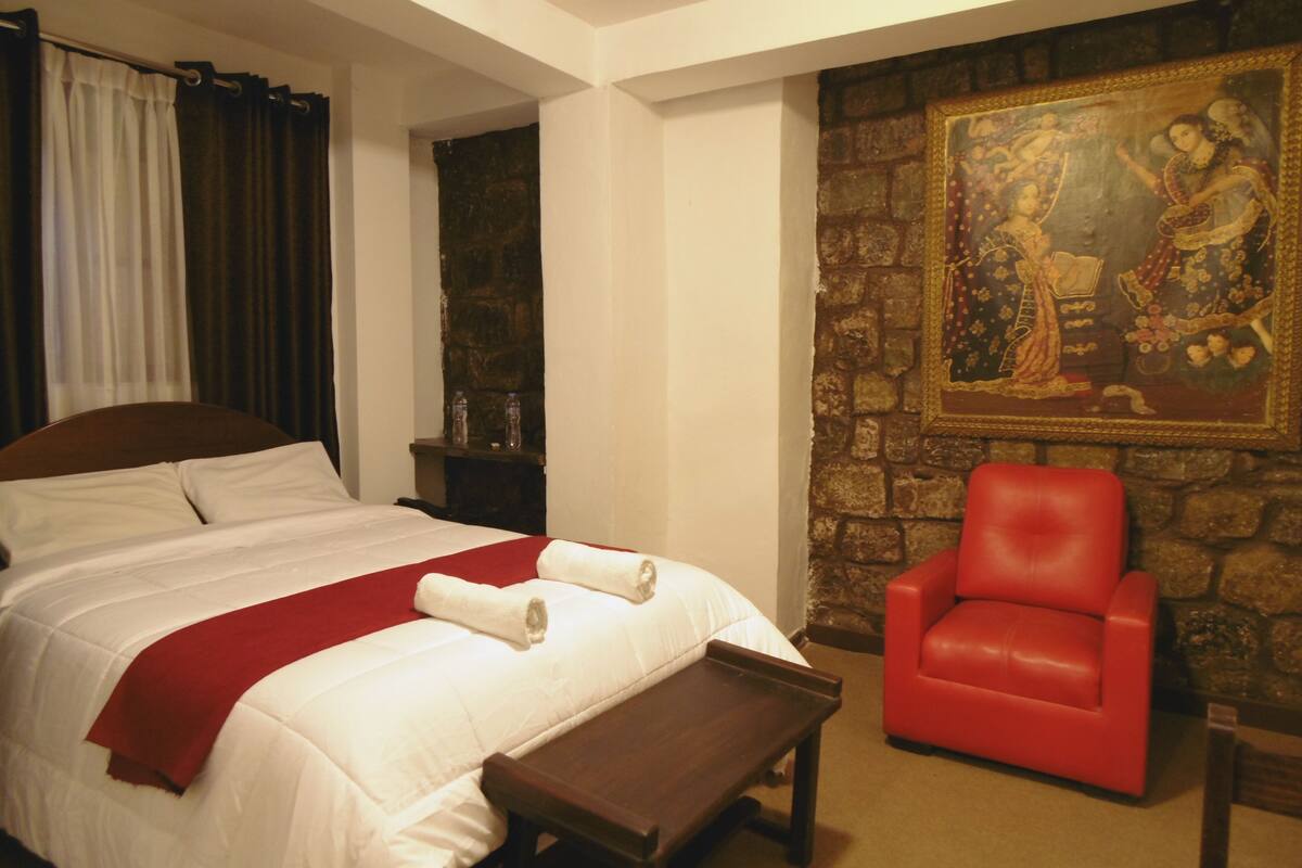 A hotel room with a large bed, a red armchair, a stone wall, and a painting.