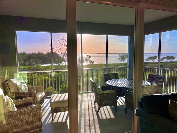 A balcony with wicker furniture, a table, and a view of a body of water and trees. - Sunset Escape - Incredible Waterfront Condo Near Beautiful Anna Maria Island! (Bradenton)