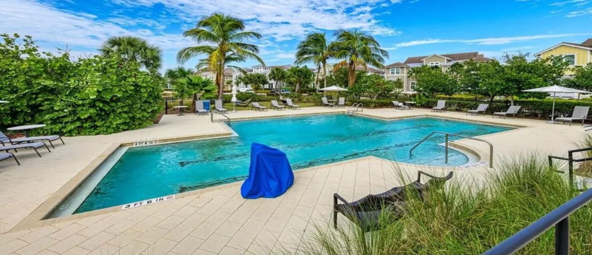 Une piscine rectangulaire recouverte d’une bâche bleue, entourée de chaises longues et de palmiers.