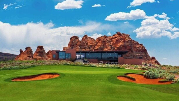 En modern byggnad med en fasad av glas, omgiven av en välskött golfbana och röda klippformationer.