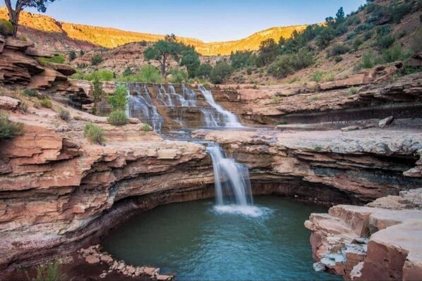 A natural waterfall cascading into a pool surrounded by rocky terrain and sparse vegetation.