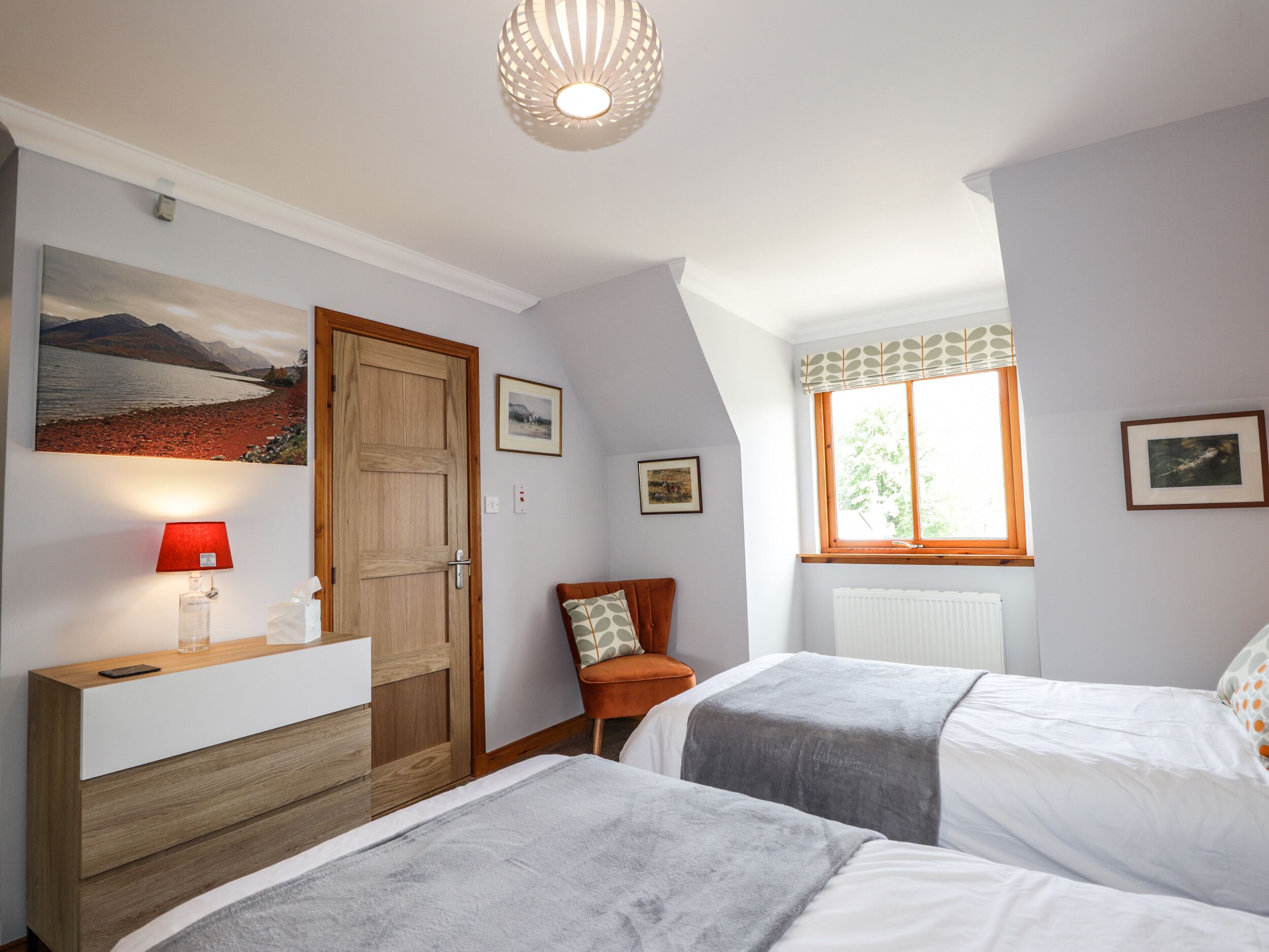 A bedroom with two beds, a wooden dresser, a red lamp, and a window with a view of the outdoors.
