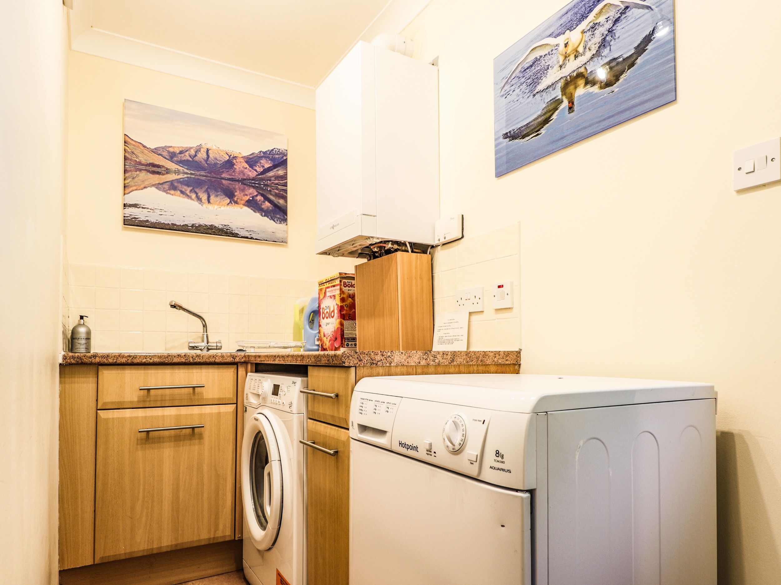 A compact kitchen with wooden cabinets, a washing machine, and a painting of a mountain landscape.