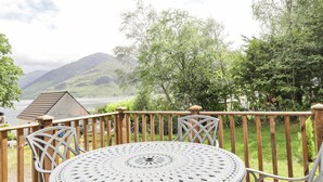Een ronde metalen tafel met een spijbelbovenblad, omringd door houten stoelen, op een balkon met uitzicht op een meer en bergen.