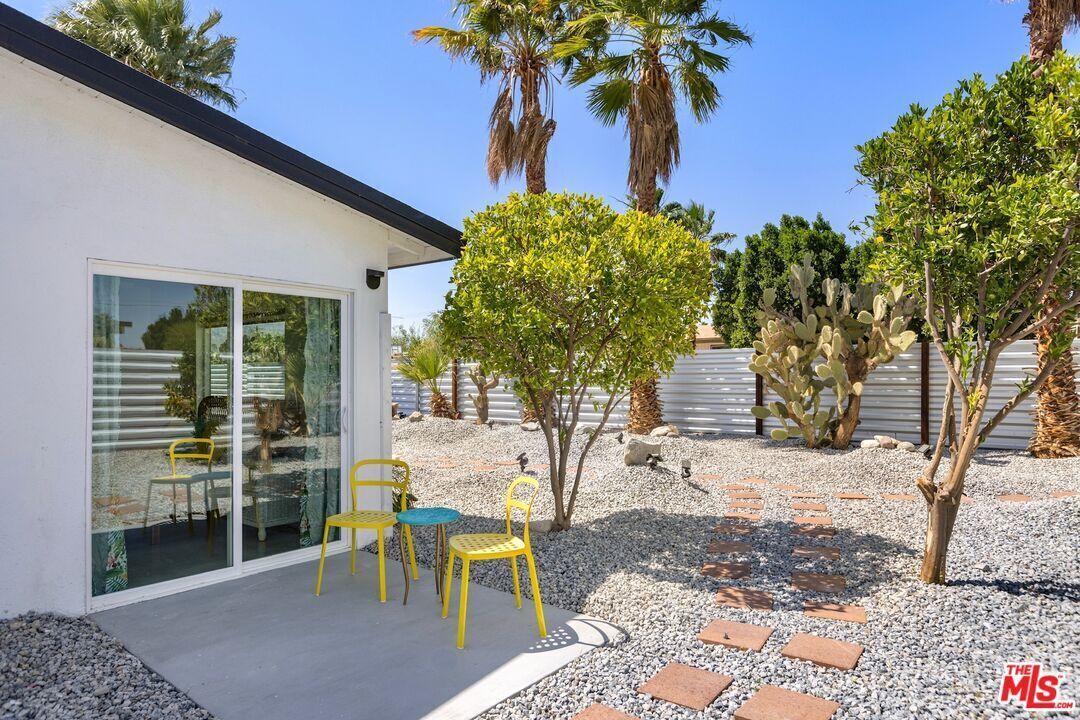 A modern house with a glass door entrance, outdoor seating with yellow chairs, and a garden with cacti and palm trees.