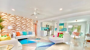 A modern living room with a sofa, coffee table, and a patterned wall. There is a kitchen area with cabinets and appliances visible through an open doorway.