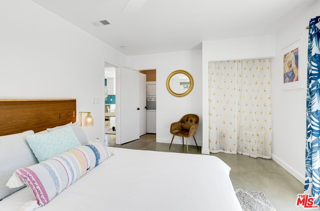 A modern bedroom with a large bed, a wooden headboard, a round mirror, a chair, and a view into the kitchen.
