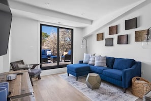 Una sala de estar moderna con un sofá seccional azul, una mesa de centro de madera y un ventanal con vistas a muebles de exterior.