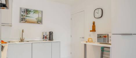 A modern kitchen with a white countertop, a sink, a coffee maker, a clock, and a small window with a beach view.