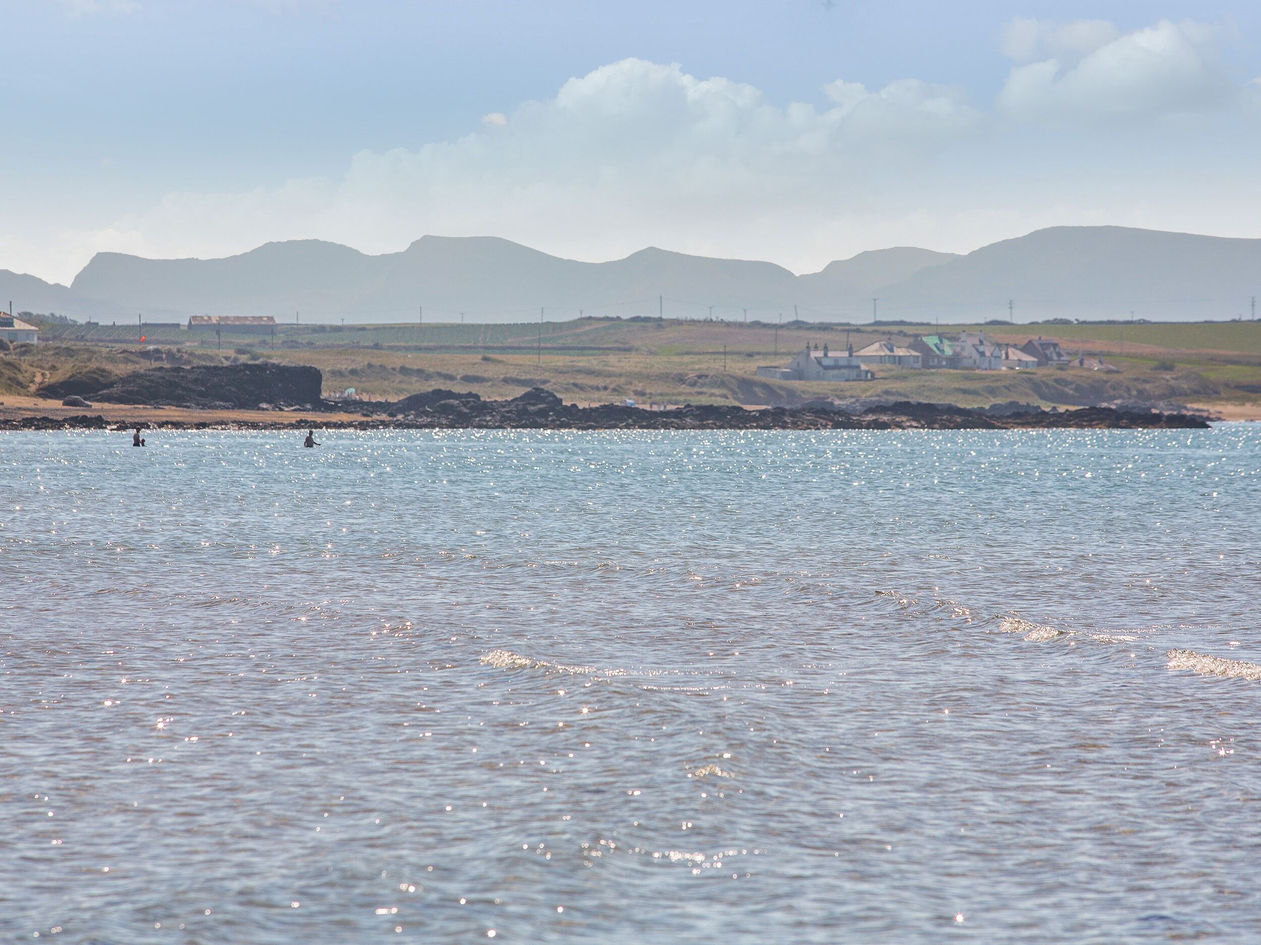 A coastal landscape with calm water, distant mountains, and a few buildings along the shore.