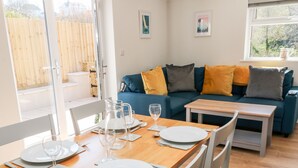 Un rincón de comedor con una mesa de madera preparada para dos, un sofá azul con cojines y una ventana con vista a follaje.