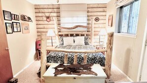 Een slaapkamer met een houten hoofdbord, een plafondventilator, een bed met beddengoed met een patroon, een nachtkastje met een lamp en een raam met uitzicht.