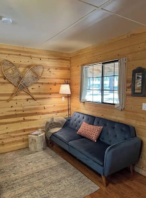 Un acogedor interior de cabaña con un sofá azul, una pared de madera, una ventana con cortinas, una lámpara y una decoración con forma de corazón.