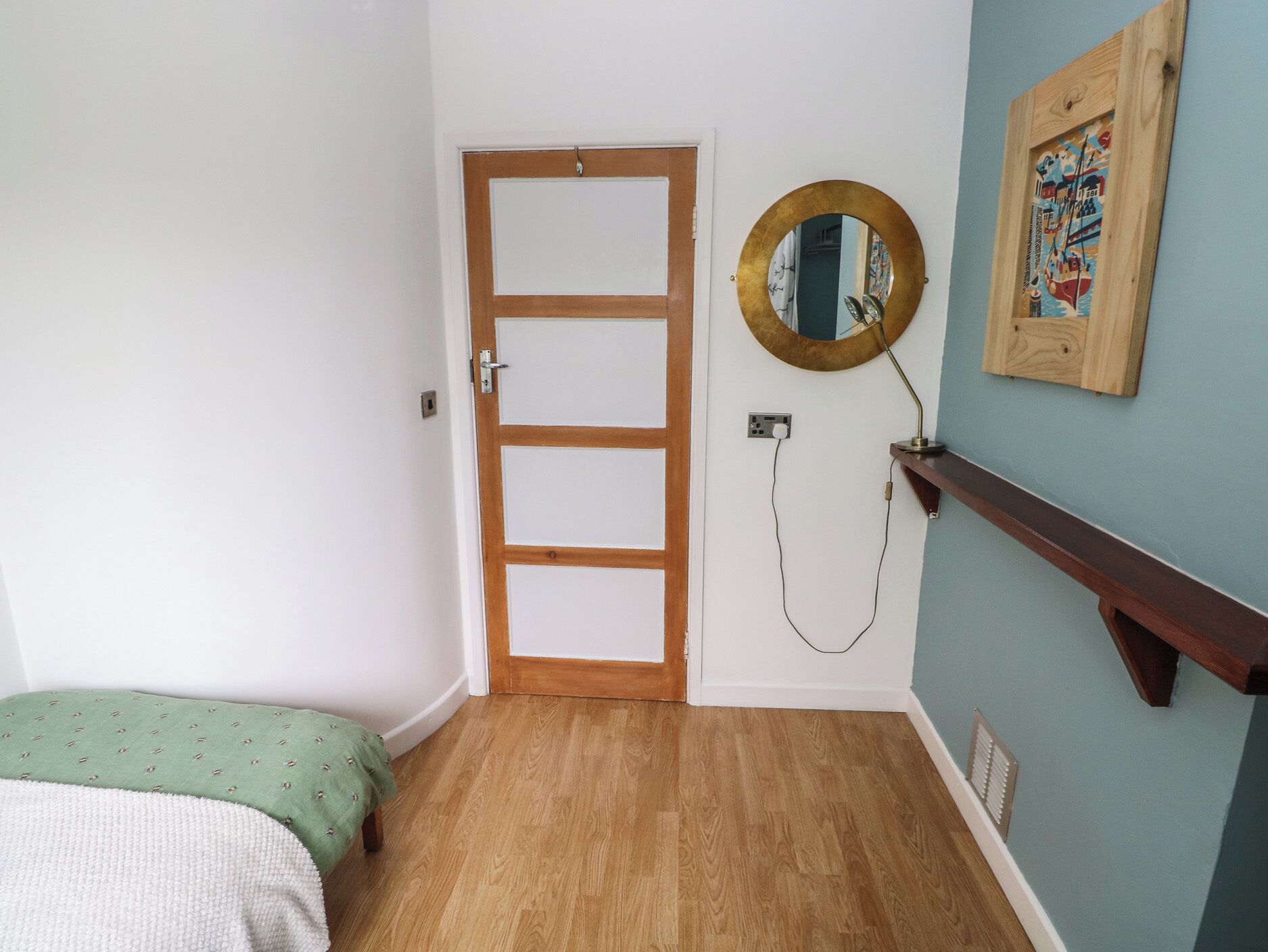 Una habitación con puerta de madera, un espejo redondo, una cama con colcha verde y blanca estampada y una estantería de madera.
