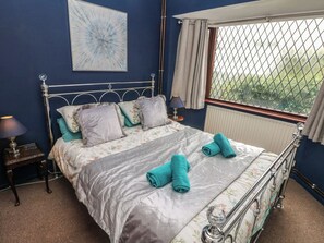 A bedroom with a metal-framed bed, floral bedding, two rolled towels, a nightstand with a lamp, and a window with a view. - Llys-yr-onnen (Carmarthen)