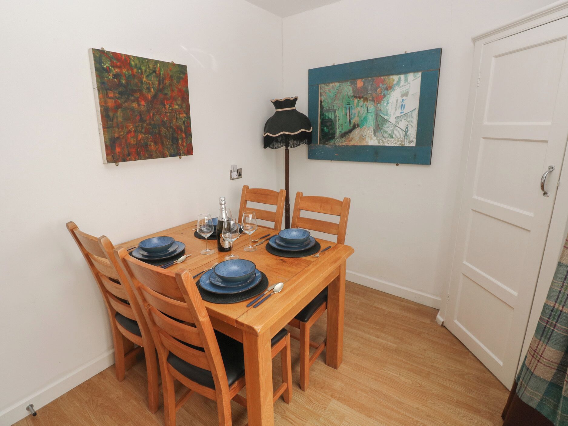 Un comedor con una mesa de madera dispuesta para cuatro personas, rodeada de sillas. Hay dos cuadros en las paredes, una lámpara de pie y una puerta con manija.