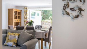 A dining area with a table set for four, a sofa with a floral cushion, and a wooden cabinet with glassware. - Stop Tide (Port Isaac)