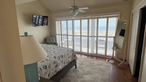 Een slaapkamer met een bed, een plafondventilator, een groot raam met jaloezieën en uitzicht op de omgeving.