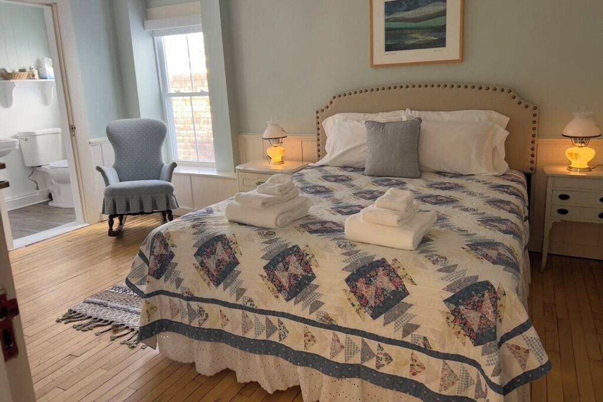 A bedroom with a bed, two bedside lamps, a chair, a window, and a painting on the wall.