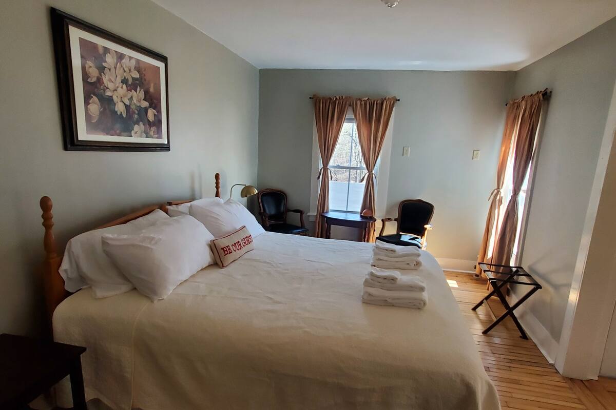 A neatly made bed with a pillow labeled "BE OUR GUESTS" in a room with a wooden floor, a framed picture on the wall, and a desk with a chair.