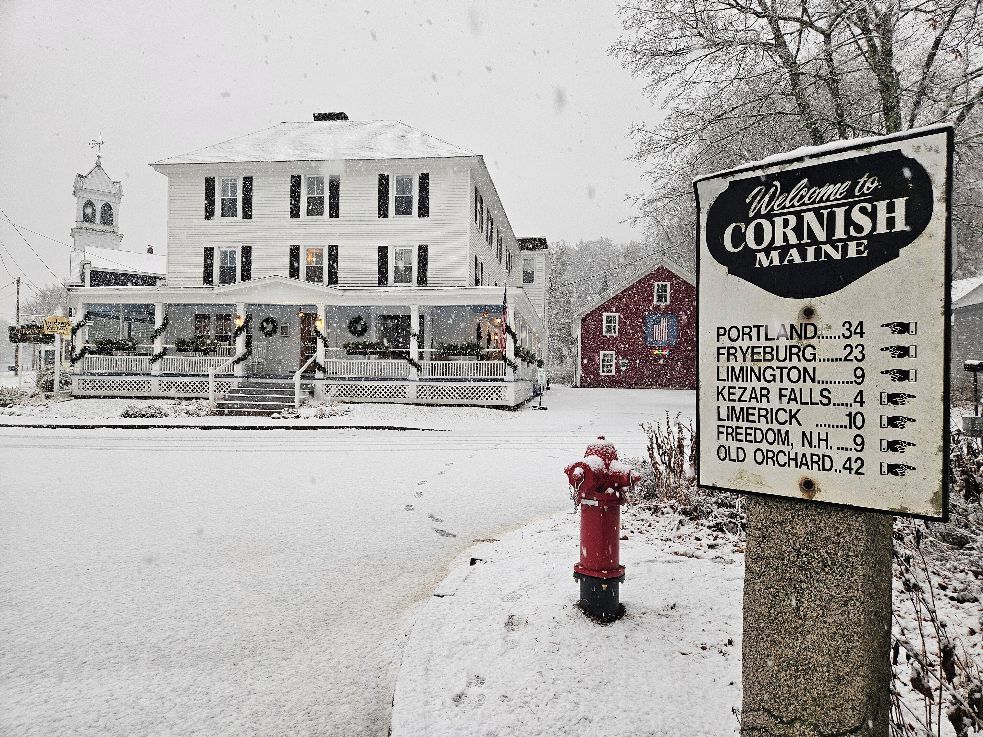 A snow-covered scene with a sign welcoming to Cornish, Maine, listing nearby towns and their distances.