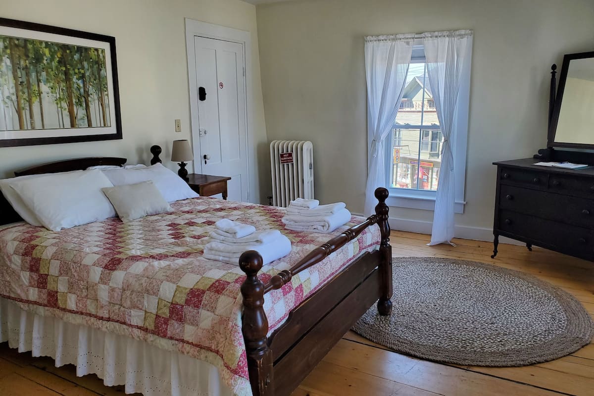A neatly made bed with a quilt, a wooden headboard, a bedside table with a lamp, a radiator, a window with curtains, and a dresser.