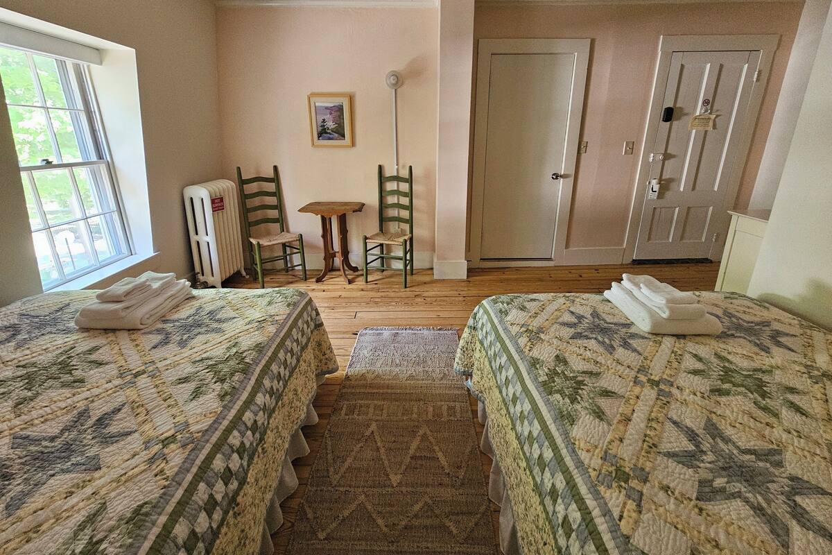 A room with two beds, a radiator, a small table, two chairs, a door, and a window.